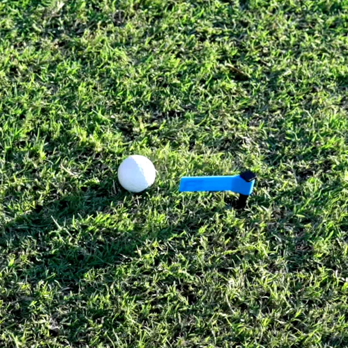 Seen from above, a FlagSwing staked in the grass just beyond a golf ball, its blue flag pointing back at the ball.